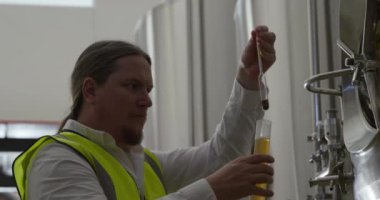 Kafkasyalı bir adam mikro bira fabrikasında çalışıyor, yüksek görüş mesafesi olan bir yelek giyiyor, bir bardak birayı inceliyor, rengini yavaş çekimde kontrol ediyor..