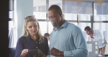 Afrika kökenli Amerikalı bir erkek ve Kafkas bir kadın iş adamının gelişigüzel modern bir ofiste tablet kullanarak ve çalışmalarını tartışarak yavaş çekimde arka planda iş arkadaşlarıyla çalışmalarını ön planda tutuyorlar.