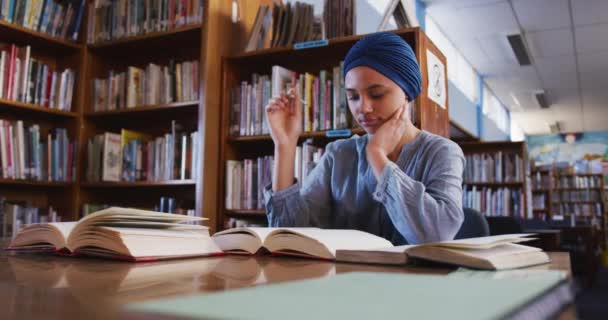 一名亚洲女生穿着蓝色头巾在图书馆学习 坐在书桌前 翻开书 做笔记 穿着休闲装 动作缓慢 — 图库视频影像
