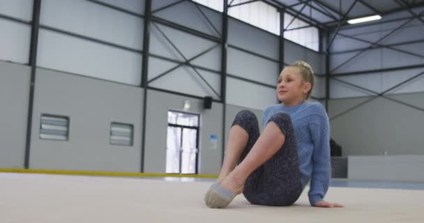 一个年轻的白人女体操运动员在体育馆练习 坐在地板上 伸展腿 体操训练对慢动作比赛很有帮助 — 图库视频影像
