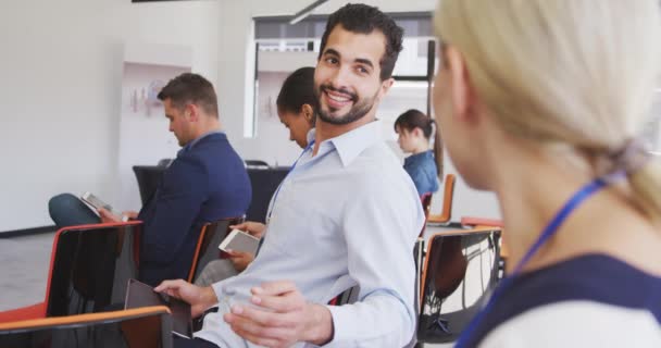 Vue de face d'un homme d'affaires mixte créatif assis dans le public lors d'une conférence d'affaires, parlant à sa collègue blanche, avec des collègues assis en arrière-plan au ralenti 