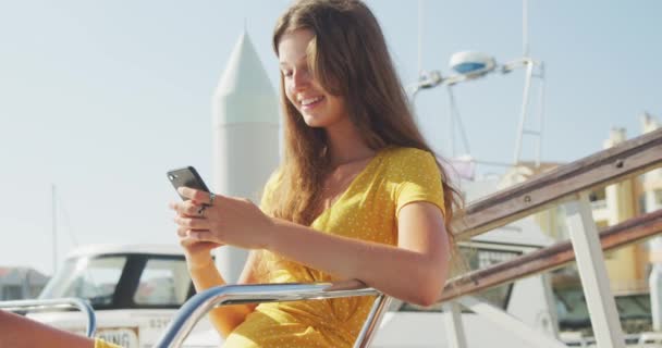 一个白人少女的侧视图 在阳光灿烂的日子里 慢慢地坐在船上 用智能手机 — 图库视频影像