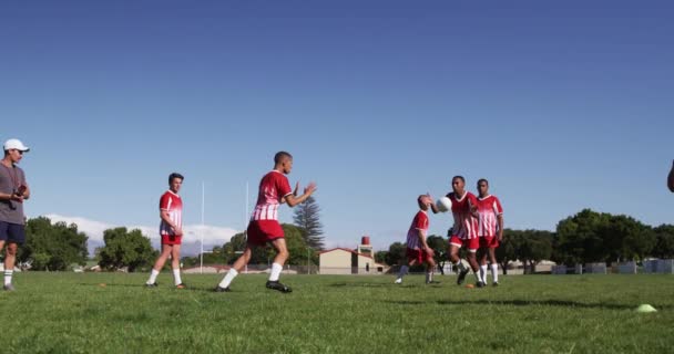 少年多种族男子橄榄球队的侧视图 橄榄球队身穿红白相间的队服 在运动场上训练 慢动作地奔跑和投掷橄榄球 — 图库视频影像