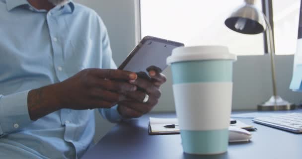 アフリカ系アメリカ人の男性のビジネスの創造的な仕事のサイドビューの真ん中のセクションでは カジュアルな近代的なオフィスでは 遅い動きで彼のスマートフォンを使用して机に座って — ストック動画
