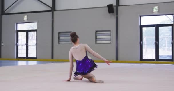 一位专注的年轻白种人女体操运动员在体育馆表演 坐在地板上 伸展胳膊 身穿紫色和白色紧身衣 动作缓慢的前景 — 图库视频影像