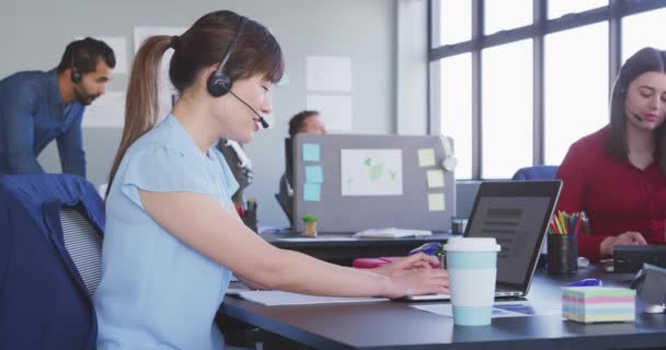 一位亚洲女商人在休闲的现代化办公室里工作 坐在办公桌前使用笔记本电脑 头戴手机耳机 而同事们则在幕后慢吞吞地工作 — 图库视频影像