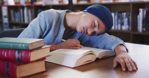 一名亚洲女生穿着蓝色头巾在图书馆学习 坐在书桌边看书 拿着铅笔睡觉 穿着休闲装 动作缓慢 — 图库视频影像