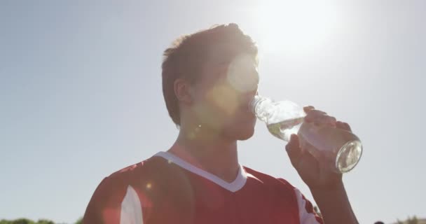 一名年轻的白种人男子橄榄球队队员穿着红白相间的队服 站在运动场上 慢动作喝水 低角度侧视 — 图库视频影像