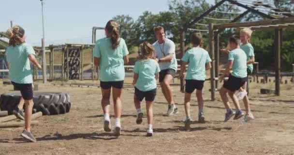 阳光灿烂的日子里 一群快乐的白人男孩女孩和一个白人男子教练在新兵训练营一起做运动 跳跃和触摸膝盖 都穿着绿色T恤 动作缓慢 — 图库视频影像