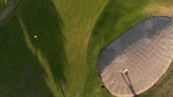 在阳光灿烂的日子里 在高尔夫球场上顶着一顶帽子 站在沙坑里 挥动高尔夫球杆 用黄色旗杆将球打出沙坑的男性高尔夫球手无人驾驶飞机向洞口射击 — 图库视频影像