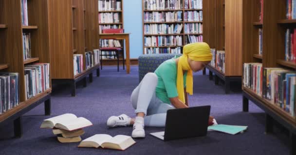 一名亚洲女学生穿着黄色头巾在图书馆学习 坐在地板上 打开书本 使用笔记本电脑 穿着休闲装 — 图库视频影像