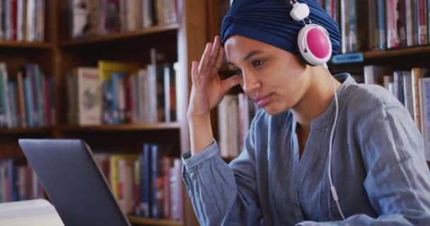 一名亚洲女生穿着蓝色头巾在图书馆学习 坐在那里使用笔记本电脑 穿着休闲装 头戴耳机 动作缓慢 — 图库视频影像