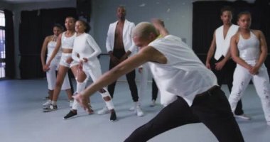 Parlak bir stüdyoda dans kursunda dans ederken beyaz kıyafetli modern erkek dansçı, takım arkadaşları arkasındayken, ağır çekimde.