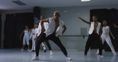Parlak bir stüdyoda dans kursunda dans ederken beyaz kıyafetli modern erkek dansçı, takım arkadaşları arkasındayken, ağır çekimde.