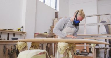 Beyaz erkek sörf tahtası yapımcısı, uzun sarı saçlı, yüz maskesi takan, stüdyosunda çalışan, sörf tahtasını yavaş çekimde cilalayan.