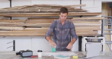 Stüdyoda çalışan beyaz erkek sörf tahtası üreticisi, tezgahın arkasında duruyor ve arka planda ağır çekimde asılı ahşap şeritlerle bir sörf tahtası projesi hazırlıyor.