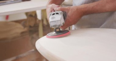 Erkek sörf tahtası yapımcısı stüdyosunda çalışıyor koruyucu bir önlük giyiyor, kum makinesiyle ahşap bir sörf tahtasını şekillendiriyor.