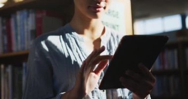 Asyalı bir kız öğrenci mavi başörtüsü takıyor, kütüphanede çalışıyor, ayakta duruyor ve tablet kullanıyor, yavaş çekimde günlük kıyafetler giyiyor.