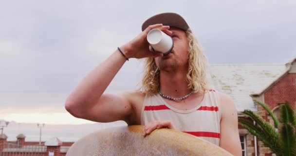 長いブロンドの髪の白人男性サーファー フルキャップとカジュアルな服を着て 木製のサーフボードを保持 スローモーションでコーヒーを飲む — ストック動画