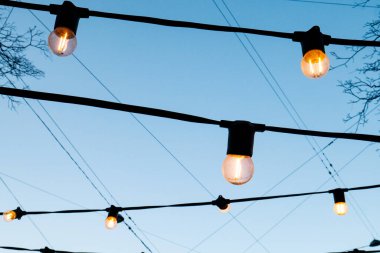 Akşam mavisi gökyüzüne karşı ampuller. Hoş, sıcak, sarı bir ışıkla parlayan fenerler..