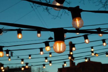 Akşam mavisi gökyüzüne karşı ampuller. Hoş, sıcak, sarı bir ışıkla parlayan fenerler..