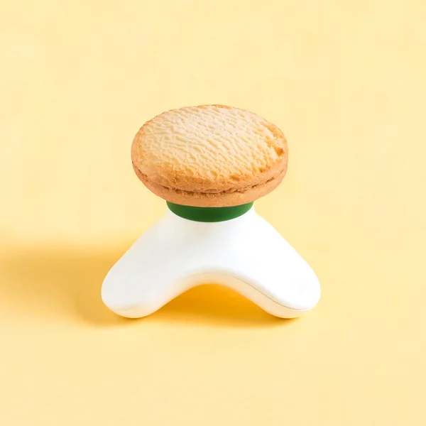 Compact massager and shortbread on pastel yellow background. Minimalist style. Creative idea, imagination and fantasy. Attraction of attention to problems of physical inactivity and abuse of sweet