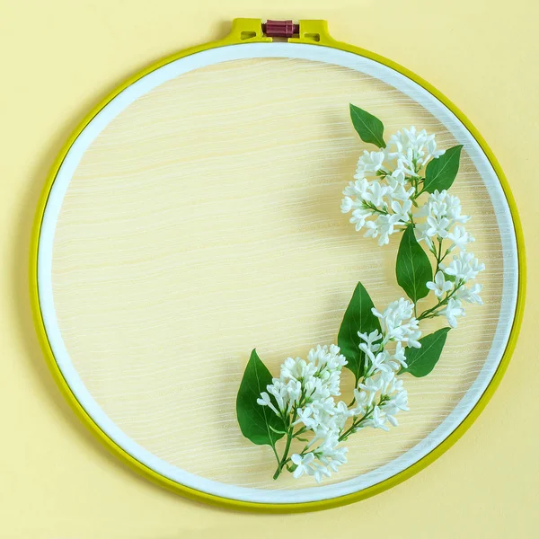 Embroidery frame and flowers of white lilac on pastel yellow background. Creative festive frame with copy space. Top view