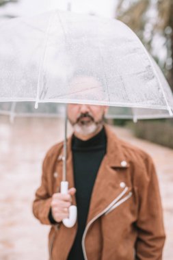 Adam yağmurda yüzünü şeffaf bir şemsiyeyle örtüyor.