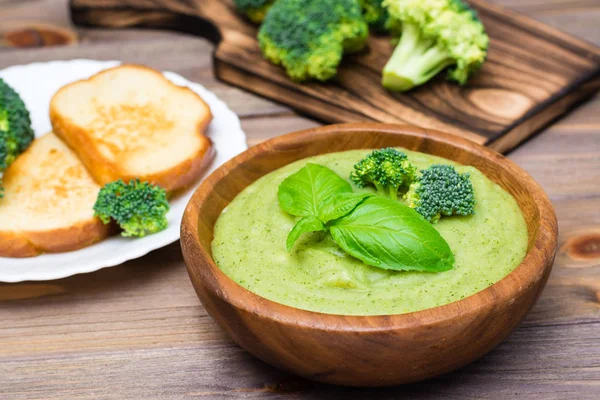 Ready to eat fresh hot broccoli puree soup with slices of broccoli and basil leaves in a wooden plate and slices of toasted white bread on a wooden table. Healthy eating and lifestyle.