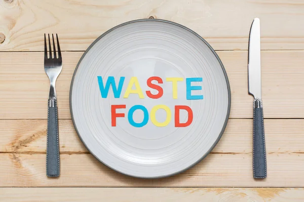 Stop wasting food. The inscription of letters cut out of colored cardboard on a plate and cutlery on a wooden background. Food waste concept