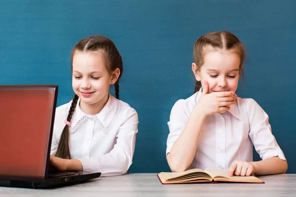 A girl works at a laptop and a girl reads a book at the table. Distance learning for children. Telework. Concept laptop versus books