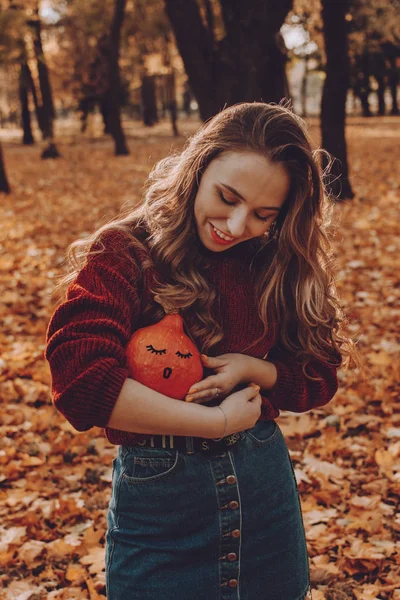 Ung vacker kvinna med långt lockigt hår håller orange halloween pumpa med ett målat ansikte på hösten park gula träd bakgrund. Höstens hösthalloween koncept — Stockfoto