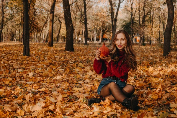 Mulher sorridente bonita com cabelos longos encaracolados sentado em folhas caídas e segurando laranja abóbora halloween no outono parque amarelo árvores fundo. Outono outono halloween conceito — Fotografia de Stock