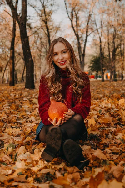 Vacker leende kvinna med långt lockigt hår sitter på fallna blad och håller orange halloween pumpa på hösten park gula träd bakgrund. Höstens hösthalloween koncept — Stockfoto