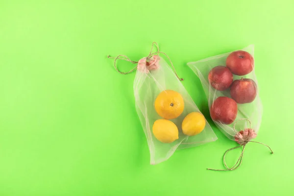 2つの再利用可能なメッシュオーガンザタートルバッグ、エコフレンドリーなパッケージ用食料品で果物、リンゴ、オレンジ、レモン緑の背景。再利用可能な生活、廃棄物ゼロ、生態系の問題とプラスチック概念 — ストック写真