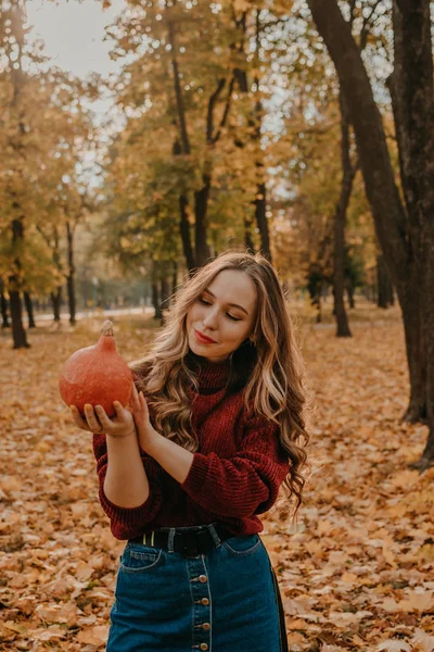 Ung vacker leende kvinna med lång lockigt hår håller orange halloween pumpa på hösten park gula träd bakgrund. Höstens hösthalloween koncept — Stockfoto