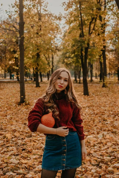 Joven hermosa mujer sonriente con pelo largo rizado sosteniendo calabaza de halloween naranja en el fondo de los árboles amarillos del parque de otoño. Otoño otoño halloween concepto —  Fotos de Stock