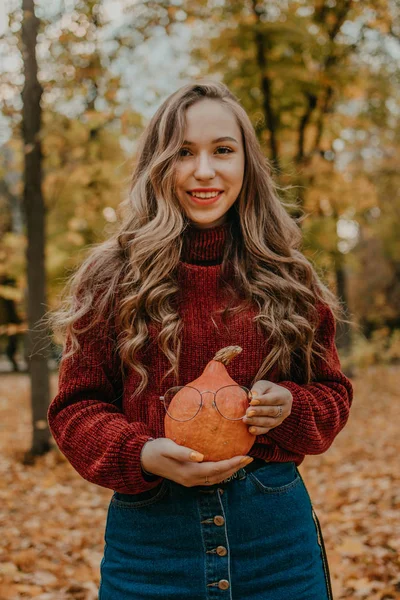 Junge schöne lächelnde Frau mit langen lockigen Haaren, die orangefarbenen Halloween-Kürbis auf herbstlichem Parkgelb hält. Herbst-Halloween-Konzept — Stockfoto