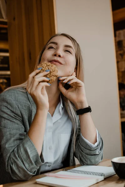 Портрет улыбающейся молодой блондинки, которая ест печенье и пьет кофе, чай в кафе. Счастливая девушка ест безглютеновое печенье — стоковое фото