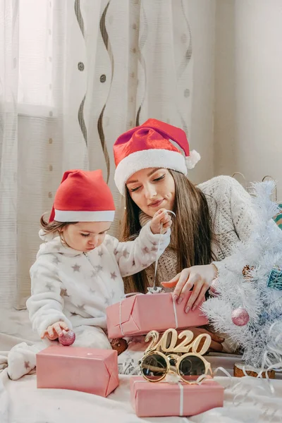 День боксу. Маленька дівчинка з мамою розпаковують рожеві коробки з подарунками біля ялинки. Хлопчик торкається яскравих різдвяних прикрас. Веселого Різдва і щасливого Нового Року. — стокове фото