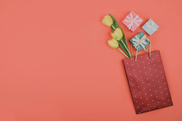 Fiori primaverili e regalo in sacchetto di carta su sfondo rosa corallo. Elegante giaciglio piatto. Concetto festivo moderno minimo . — Foto Stock