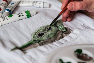 Bebek Yoda akrilik boyalarla kumaş çiziyor. Bebek Yoda, Star Wars filmlerindeki popüler karakter ve Mandalorian TV dizisi Lucasfilm hayran sanatı. Kropivnitskiy, Ukrayna, 7 Ocak 2020.