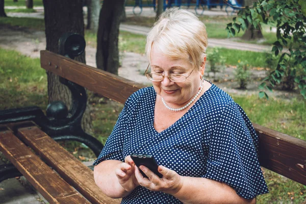 Cheerful old woman with smartphone at park. Outdoor portrait of happy senior woman at park using mobile phone. Plus size blond elderly woman have video call.