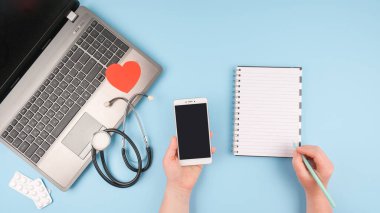 Teletıp veya tele-sağlık sanal ziyareti, video ziyareti, bilgisayarlı uzaktan doktor görüntülü sohbet konsepti, doktor eli, akıllı telefon, not defteri ve mavi arka planda steteskop.