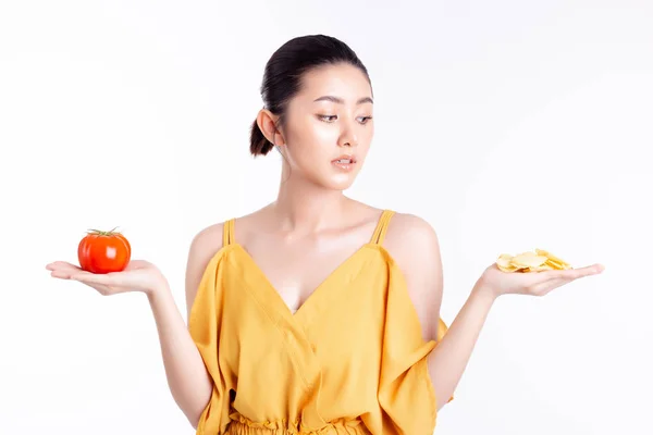 Ernährungskonzept. schöne junge asiatische Frau schaut auf leckeren Snack, Kartoffelchips. attraktive Mädchen überlegen, Zurückhaltung für das Aufhören Snack essen, weil sie Schlankheitspläne bekommt. sie halten Tomate, Kartoffelchips — Stockfoto