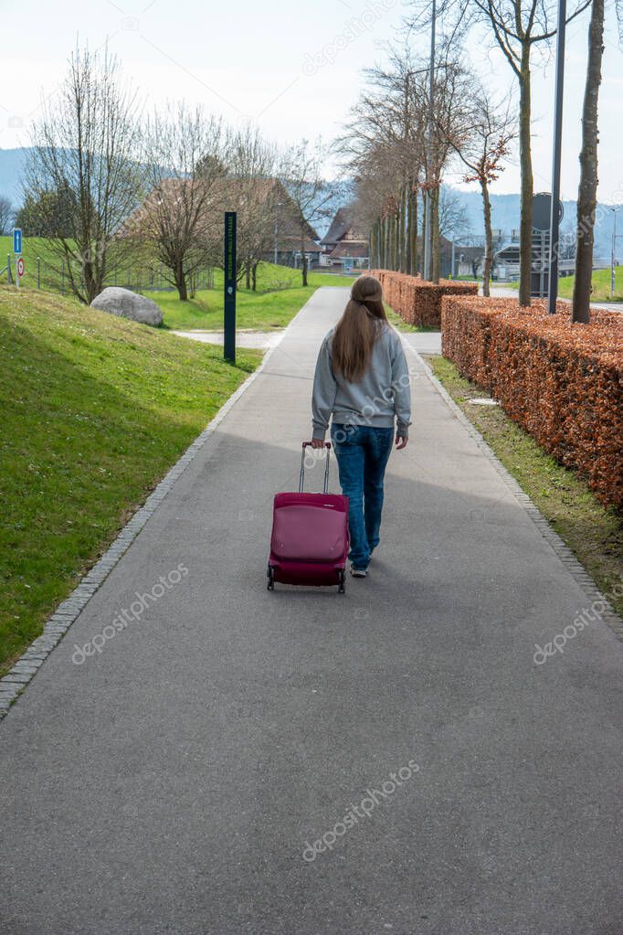 Young Caucasian woman walking on a sidewalk dragging her roller luggage case viewed from behind unrecognizable 2020