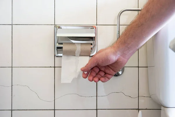 部分的に見えるトイレの洗面台と金属配管の横にほぼ空のトイレのロールのために到達白人男性の手2020 — ストック写真