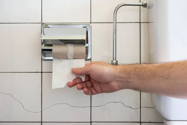 Caucasian male hand reaching for a nearly empty toilet roll next to a partially visible toilet basin and metal plumbing 2020