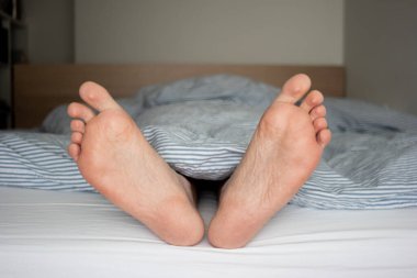 Beyaz erkek ayakları yatakta yorgandan sarkıyor. Saha manzarasının derinliği 2020.