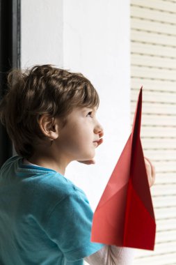 Elinde kırmızı kağıt bir uçakla pencereden bakan bir çocuğun portresi..